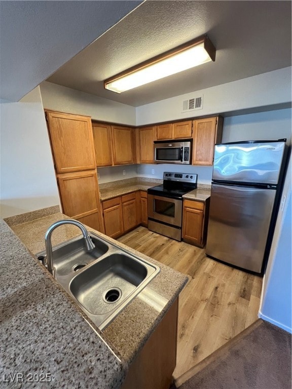 Kitchen featuring stainless steel appliances, brown cabinetry, light wood finished floors, a textured ceiling, and light stone countertops