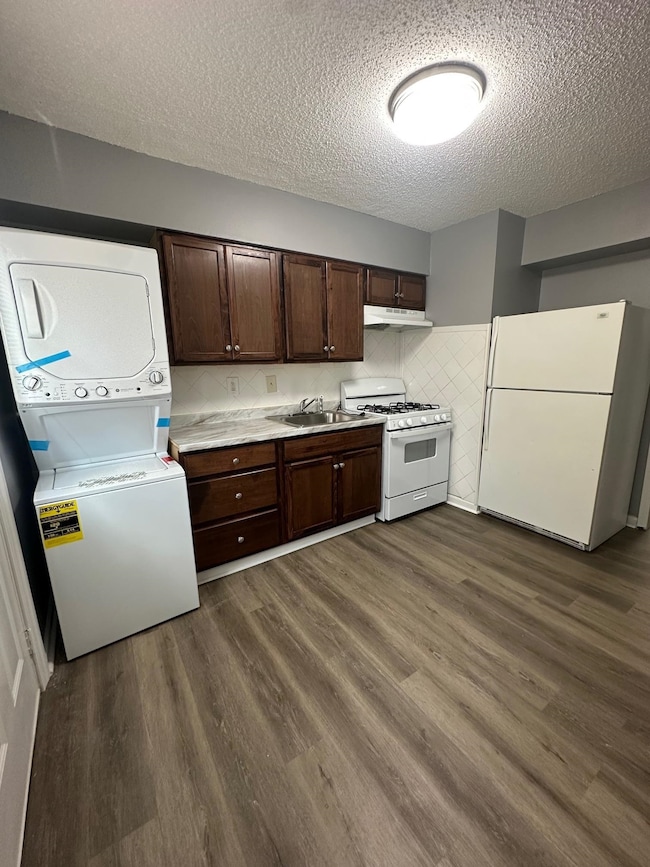 Kitchen with white appliances, estacked washer and dryer, under cabinet range hood, a textured ceiling, and dark brown cabinetry
