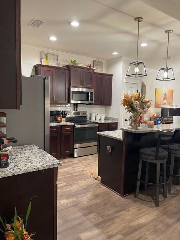 Kitchen with dark brown cabinetry, appliances with stainless steel finishes, ornamental molding, light stone counters, and hanging light fixtures