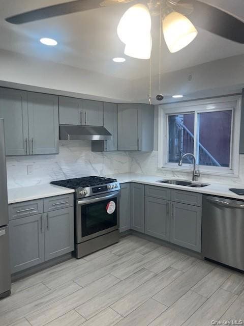 Kitchen with gray cabinets, appliances with stainless steel finishes, decorative backsplash, light stone counters, and under cabinet range hood