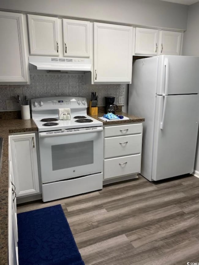 Kitchen featuring white appliances, under cabinet range hood, white cabinetry, and dark countertops