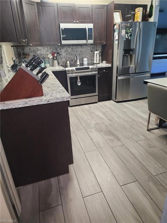 Kitchen featuring appliances with stainless steel finishes, wood tiled floors, dark brown cabinets, decorative backsplash, and light stone countertops