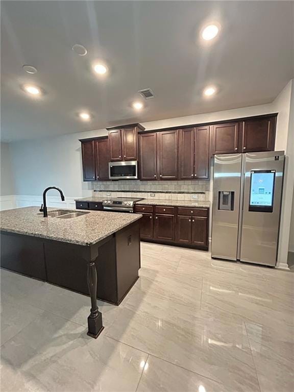 Kitchen with appliances with stainless steel finishes, light stone counters, dark brown cabinets, light marble finish flooring, and backsplash