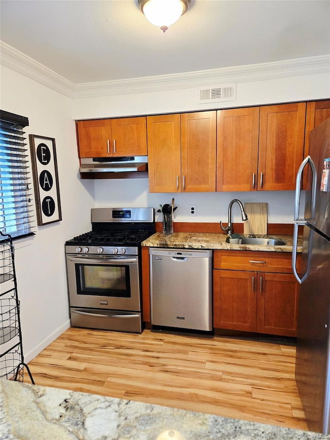 Kitchen featuring appliances with stainless steel finishes, light stone countertops, brown cabinets, crown molding, and light wood-style floors