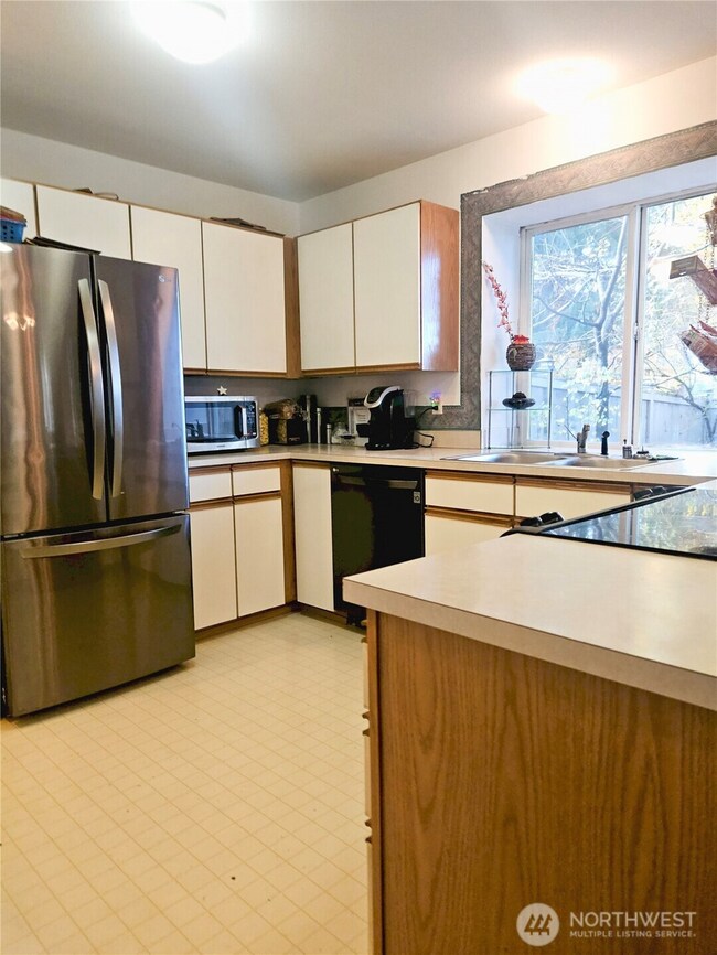 This additional view of the kitchen highlights its spacious layout, abundant cabinetry, and updated stainless-steel refrigerator. Large windows keep the space bright and inviting, creating a comfortable spot for cooking, gathering, and everyday routines.