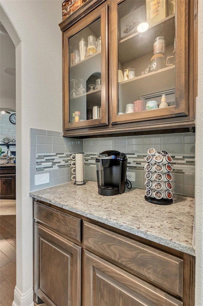 Bar with decorative backsplash, light stone counters, arched walkways, brown cabinetry, and glass insert cabinets