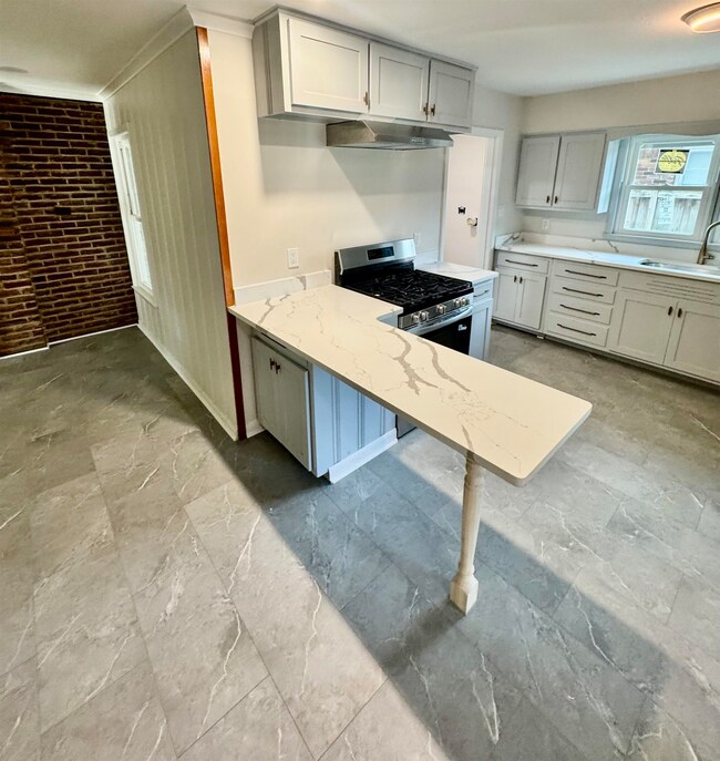 Kitchen with stainless steel range with gas cooktop, ventilation hood, light stone counters, brick wall, and a peninsula