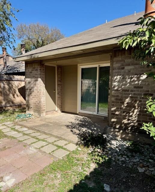 View of exterior entry with brick siding, roof with shingles, and a patio