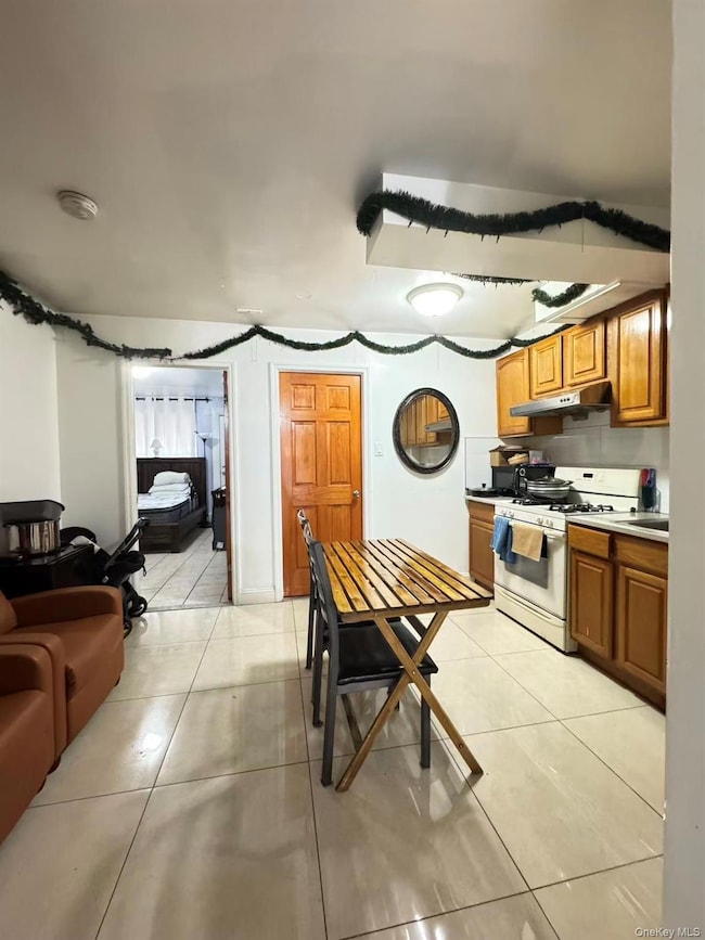 Kitchen featuring white gas stove, brown cabinets, light tile patterned floors, under cabinet range hood, and light countertops