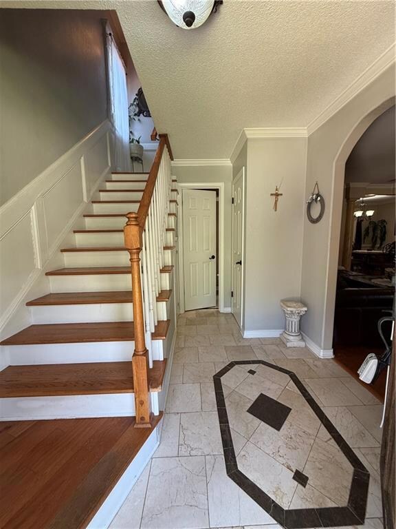 Stairway with arched walkways, crown molding, stone tile flooring, a wainscoted wall, and baseboards