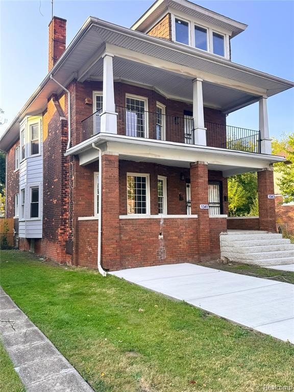 View of front of property with brick siding, a porch, a chimney, a front lawn, and a balcony