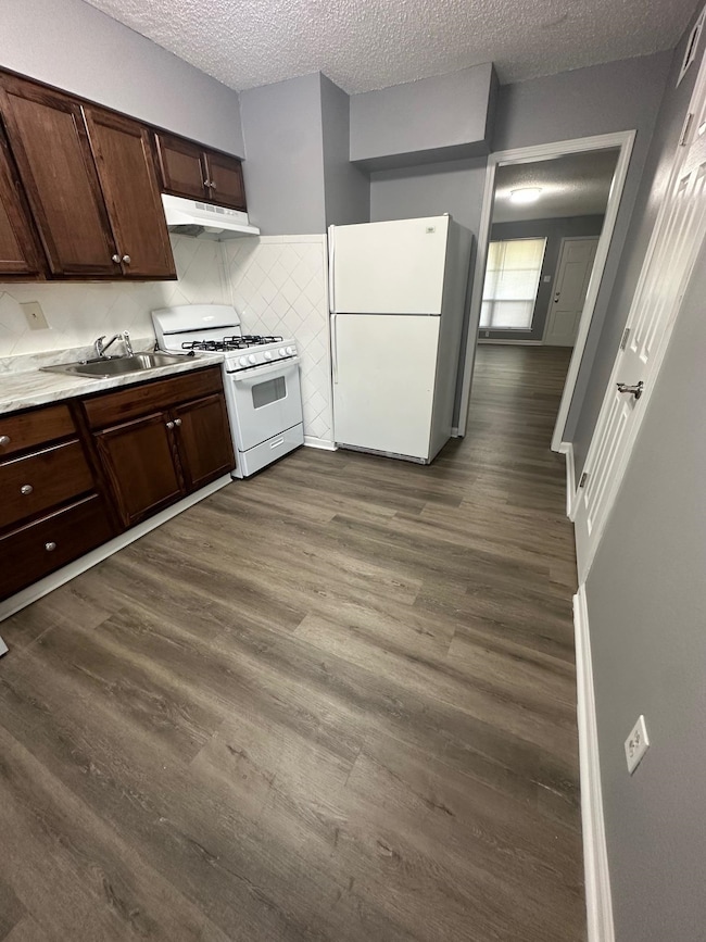 Kitchen featuring white appliances, under cabinet range hood, a sink, dark wood-style floors, and a textured ceiling