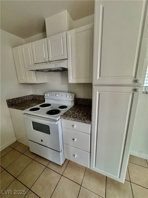 Kitchen featuring white electric range oven, light tile patterned floors, white cabinets, under cabinet range hood, and dark stone counters