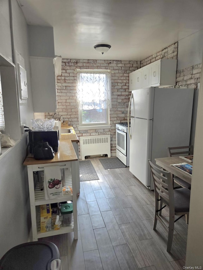 Kitchen with brick wall, white cabinetry, white appliances, radiator, and light wood-type flooring