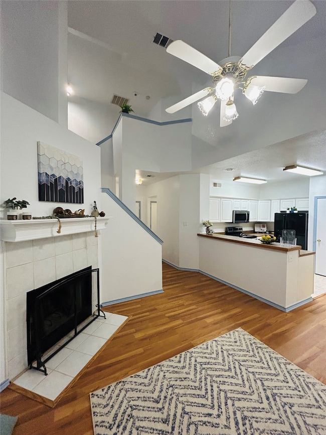 Living room featuring a tiled fireplace, light wood-type flooring, ceiling fan and a high ceiling.