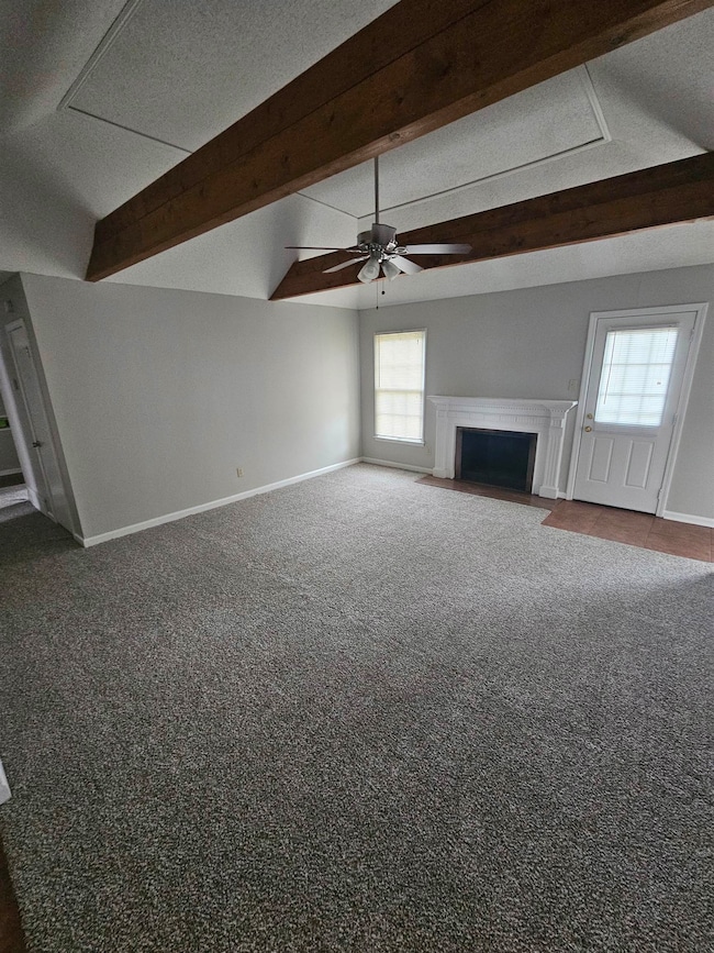 Unfurnished living room featuring a ceiling fan, beamed ceiling, baseboards, dark colored carpet, and a fireplace with flush hearth