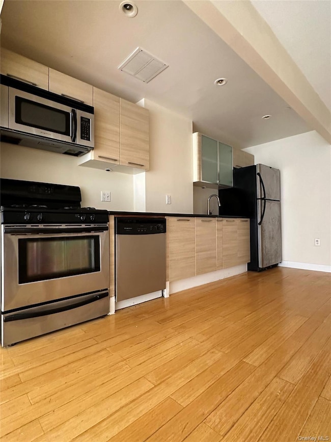 Kitchen with stainless steel appliances, light brown cabinetry, light wood-style flooring, modern cabinets, and dark countertops