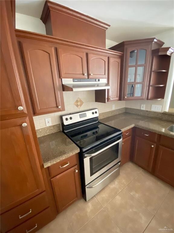 Kitchen with stainless steel range with electric cooktop, under cabinet range hood, backsplash, light tile patterned floors, and glass insert cabinets