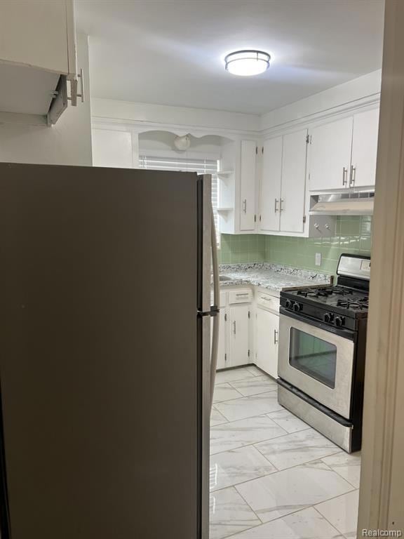 Kitchen featuring stainless steel appliances, decorative backsplash, light marble finish flooring, white cabinets, and under cabinet range hood