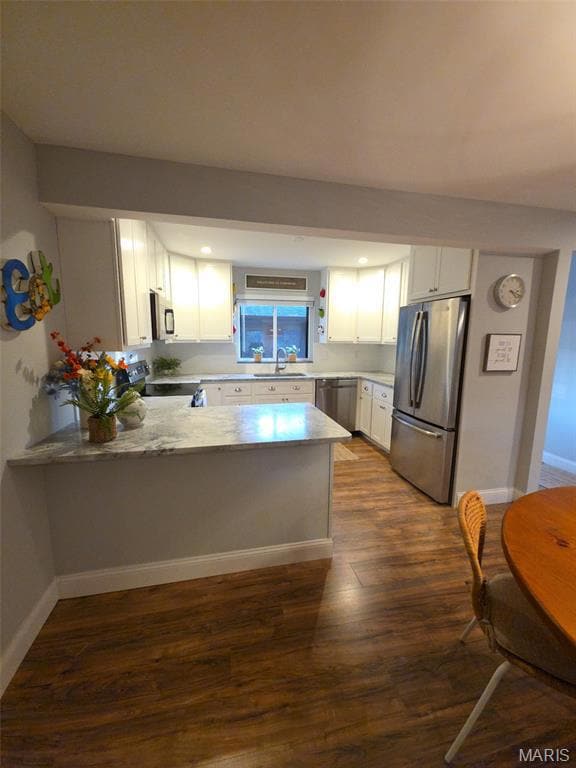 Kitchen featuring white cabinets, stainless steel appliances, a peninsula, light stone countertops, and dark wood finished floors