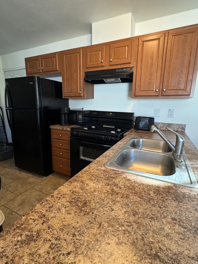 Kitchen featuring brown cabinetry, black appliances, under cabinet range hood, and light tile patterned flooring