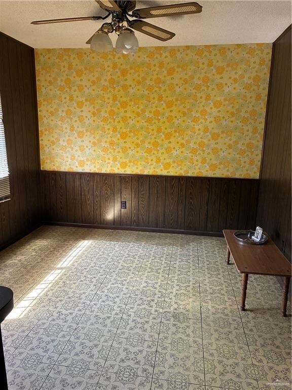 Empty room featuring wooden walls, a textured ceiling, wainscoting, ceiling fan, and wallpapered walls