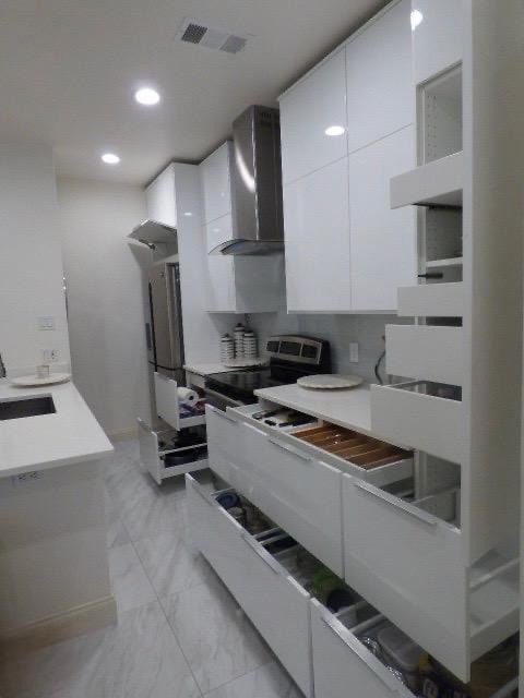 Kitchen featuring modern cabinets, white cabinetry, wall chimney range hood, light marble finish flooring, and recessed lighting