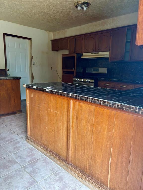 Kitchen featuring a peninsula, a textured ceiling, light tile patterned floors, stainless steel gas range oven, and tile counters