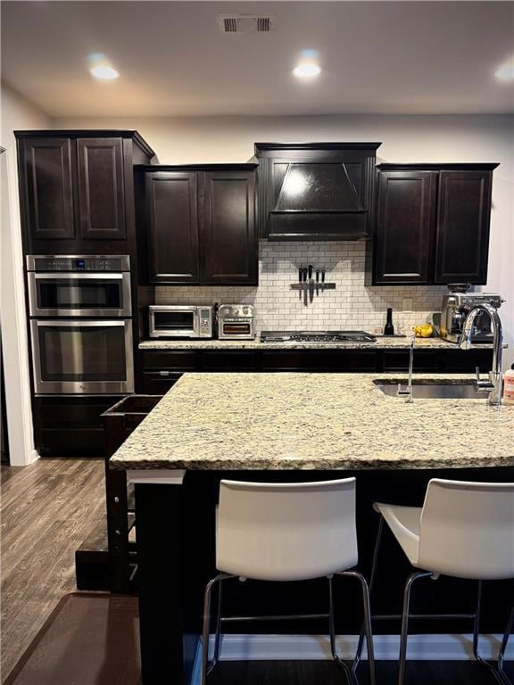 Kitchen with custom exhaust hood, stainless steel gas stovetop, a breakfast bar, light stone counters, and decorative backsplash
