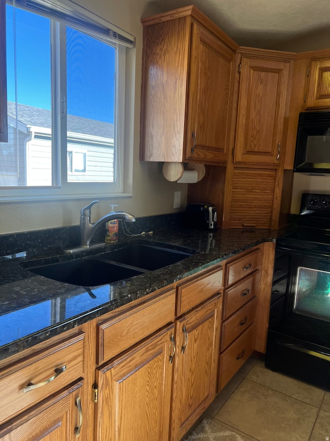 Kitchen featuring brown cabinets, black appliances, dark stone counters, and light tile patterned flooring
