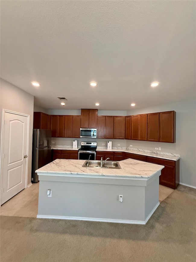 Kitchen with light colored carpet, a center island with sink, recessed lighting, stainless steel appliances, and brown cabinets