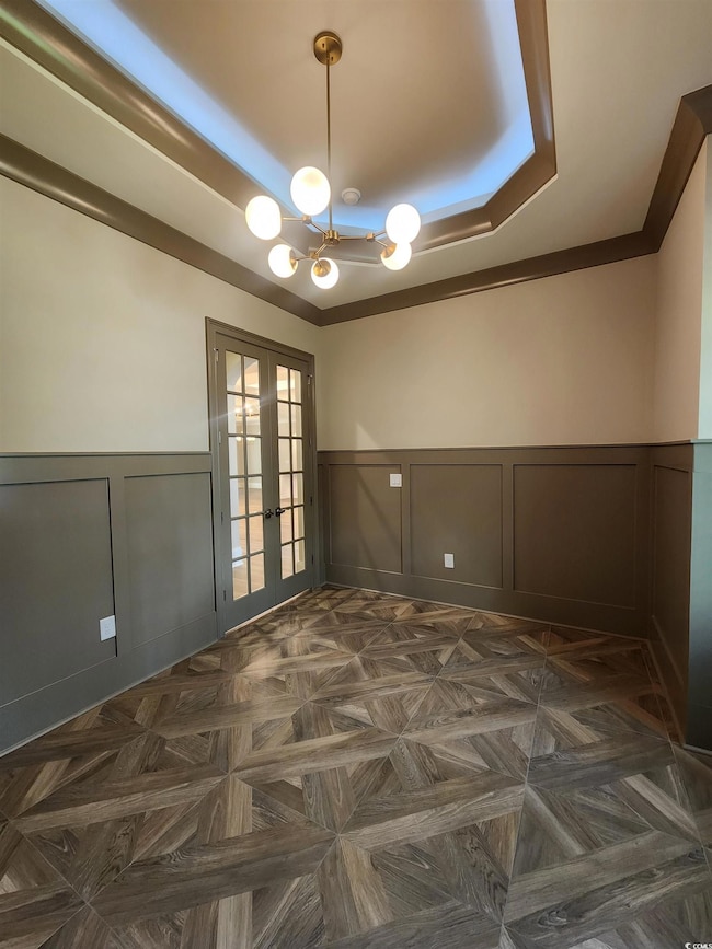 Empty room with a tray ceiling, french doors, a decorative wall, a chandelier, and wainscoting