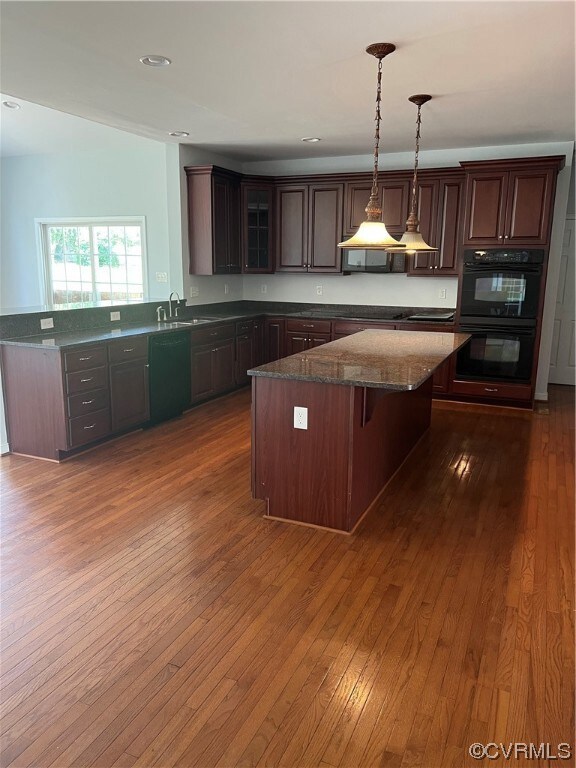 Kitchen with hanging light fixtures, double oven, a center island with sink, dark brown cabinetry, hardwood floors, dishwashing machine, and dark countertops