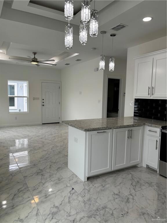 Kitchen featuring dark stone counters, ceiling fan with notable chandelier, decorative light fixtures, a raised ceiling, and white cabinetry