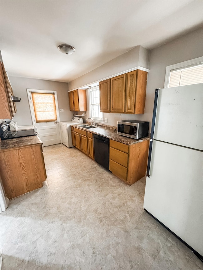 Kitchen with washer and dryer, brown cabinetry, freestanding refrigerator, black dishwasher, and stainless steel microwave