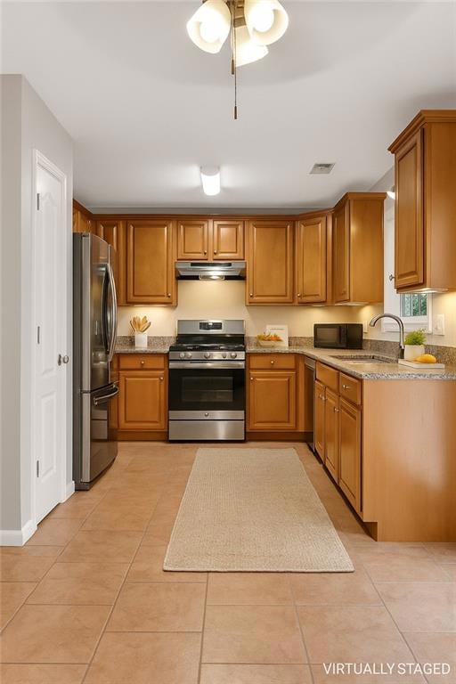 Kitchen with stainless steel appliances, under cabinet range hood, brown cabinets, light tile patterned floors, and light stone countertops