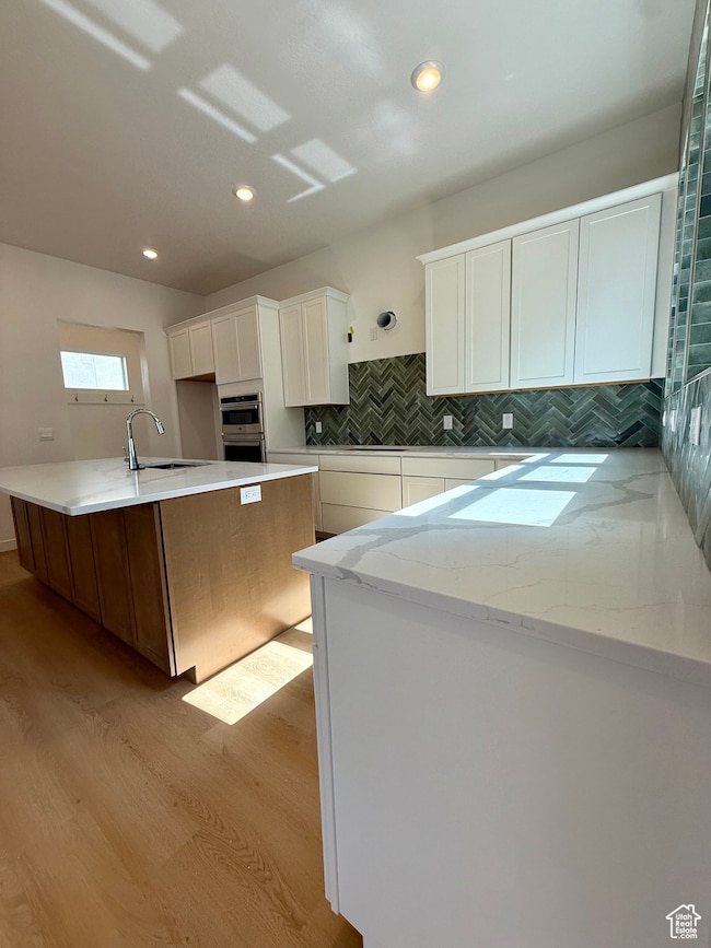Kitchen featuring white cabinetry, backsplash, light stone counters, light wood-type flooring, and an island with sink