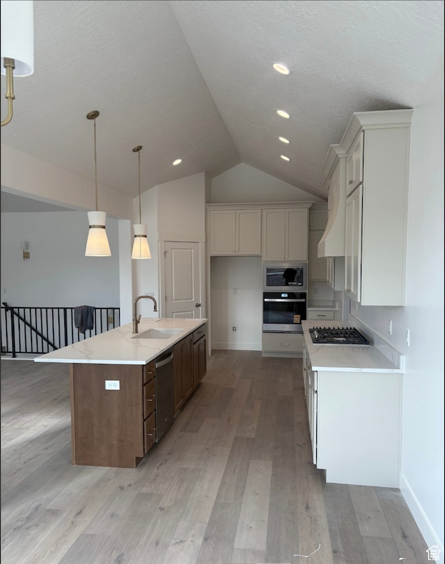 Kitchen with stainless steel appliances, lofted ceiling, an island with sink, light stone countertops, and light wood finished floors