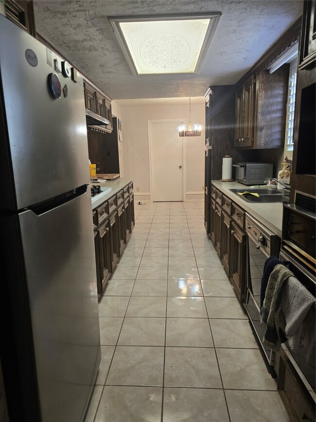 This kitchen features a long, narrow design with ample dark wood cabinetry on both sides. It has a tile floor, stainless steel refrigerator, and a stylish ceiling light fixture, offering a functional and classic look.