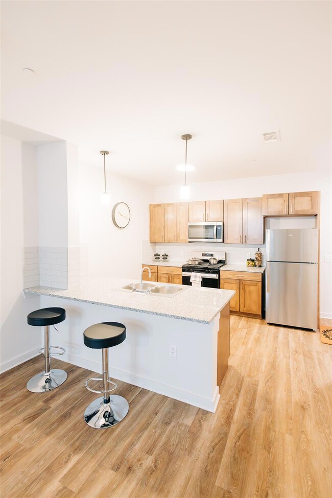 Kitchen with light brown cabinetry, stainless steel appliances, hanging light fixtures, a breakfast bar area, and light wood finished floors