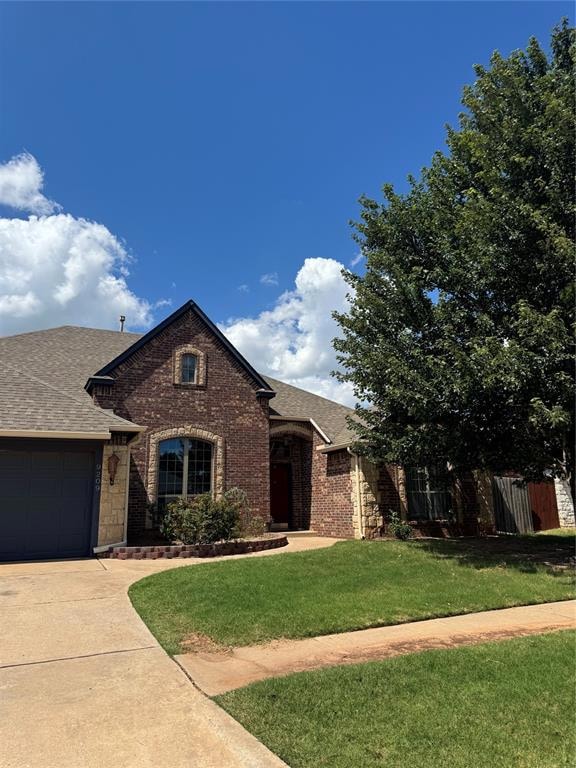 View of front of house featuring roof with shingles, driveway, brick siding, a front yard, and an attached garage