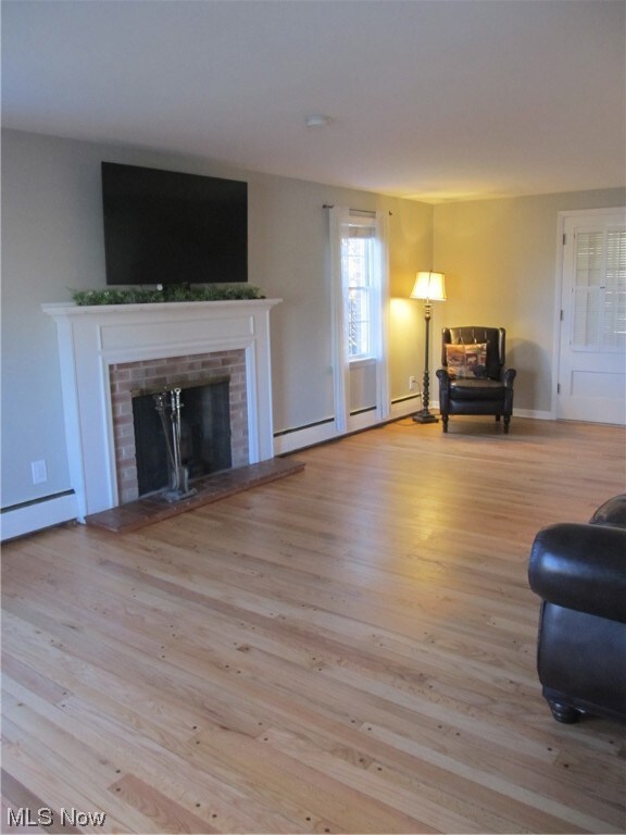 Living room with a brick woodburning fireplace, a baseboard radiator, and recently refinished hardwood floors. Door at rear of room leads to large, enclosed sun rm/porch.