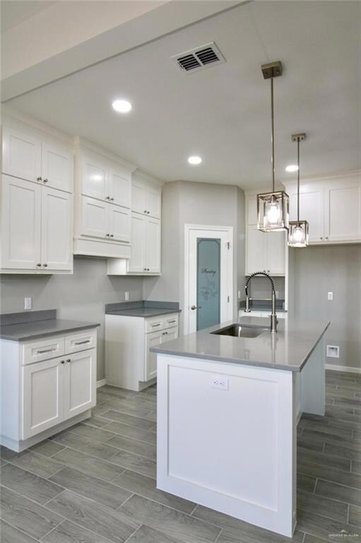 Kitchen with decorative light fixtures, wood-type flooring, sink, white cabinets, and a kitchen island with sink