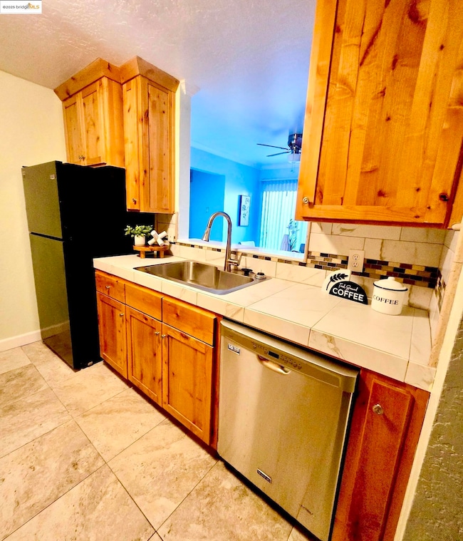 Kitchen with tile counters, freestanding refrigerator, stainless steel dishwasher, decorative backsplash, and a textured ceiling