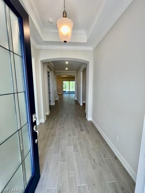Hallway featuring arched walkways, light wood-type flooring, baseboards, a raised ceiling, and ornamental molding