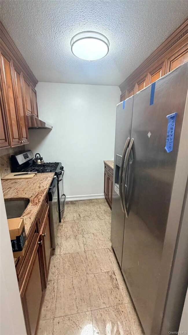 Kitchen with stainless steel appliances, light stone counters, a textured ceiling, under cabinet range hood, and brown cabinets