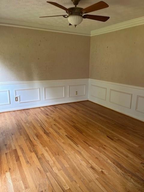 Unfurnished room featuring a ceiling fan, a wainscoted wall, ornamental molding, wood finished floors, and a decorative wall
