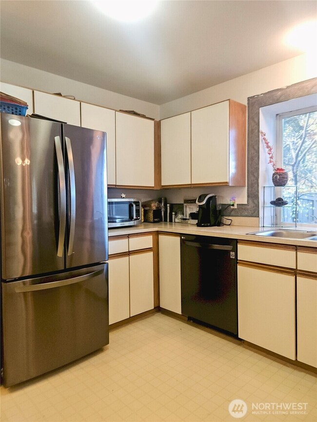 This angle showcases the roomy U-shaped layout, offering generous cabinet storage and ample counter space for cooking and coffee-making alike. The stainless-steel refrigerator and large window add both functionality and natural light, making the kitchen feel bright, practical, and inviting.