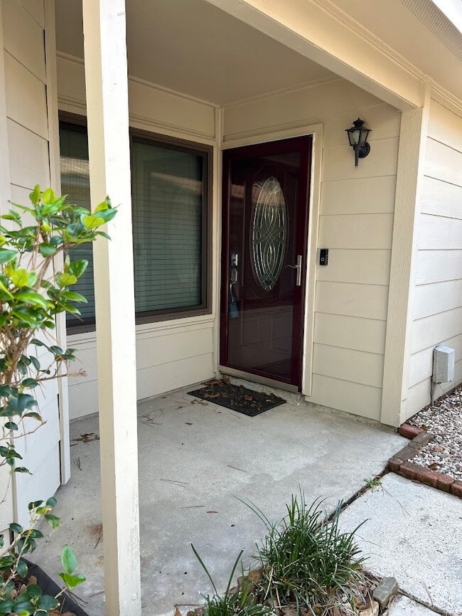 This inviting entryway features a decorative glass front door, a covered porch area, and a small garden bed adding a touch of greenery. The exterior is clean and well-maintained, with neutral siding and a wall-mounted light fixture.