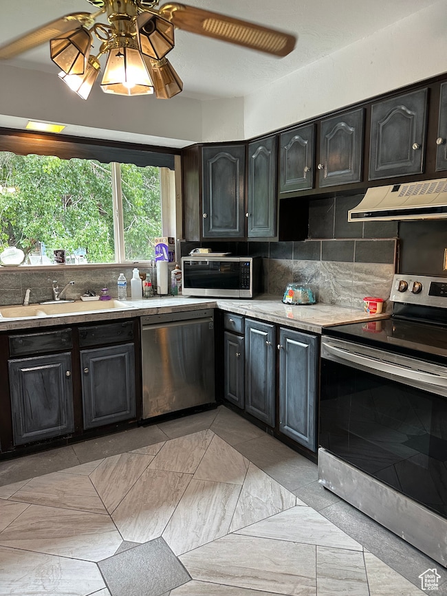 Kitchen with stainless steel appliances, backsplash, a ceiling fan, under cabinet range hood, and dark cabinets